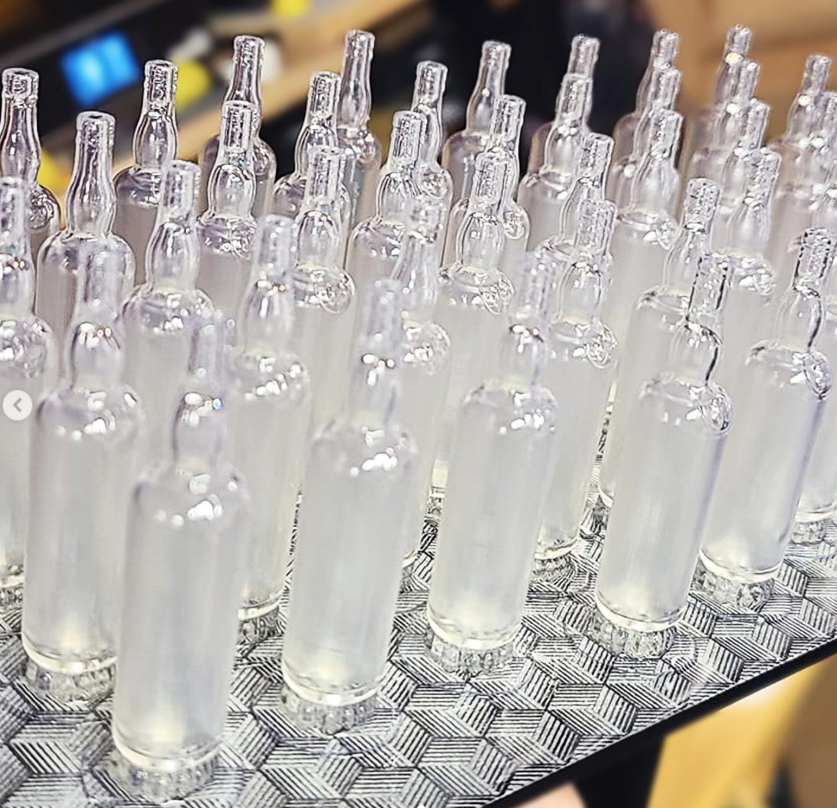 A tray of 3D-printed miniature J&B Rare Scotch Whiskey bottle replicas, made with transparent resin, featuring a smooth, glass-like finish.