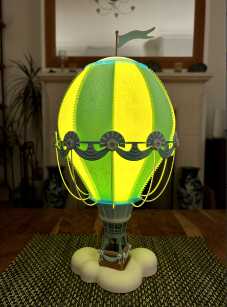 A brightly lit 3D-printed hot air balloon lamp with a yellow and green balloon, suspended over cloud-shaped base for a whimsical effect.