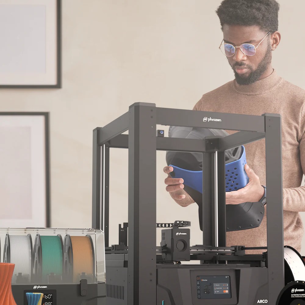 Man lifting a blue cosplay helmet from an ARCO 3D printer in a workshop.